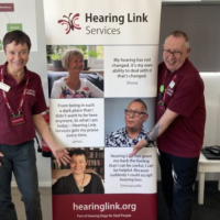 emmanuelle and james, peer support volunteers, pointing to their faces on a hearing link services roller banner