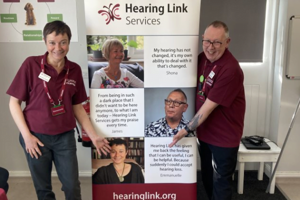 emmanuelle and james, peer support volunteers, pointing to their faces on a hearing link services roller banner